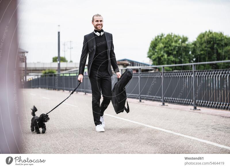 Modischer junger Mann hält Gitarrenkoffer mit Hund auf einer Brücke gehen gehend geht Bruecken Brücken modisch Hunde Männer männlich Haustier Haustiere Tier