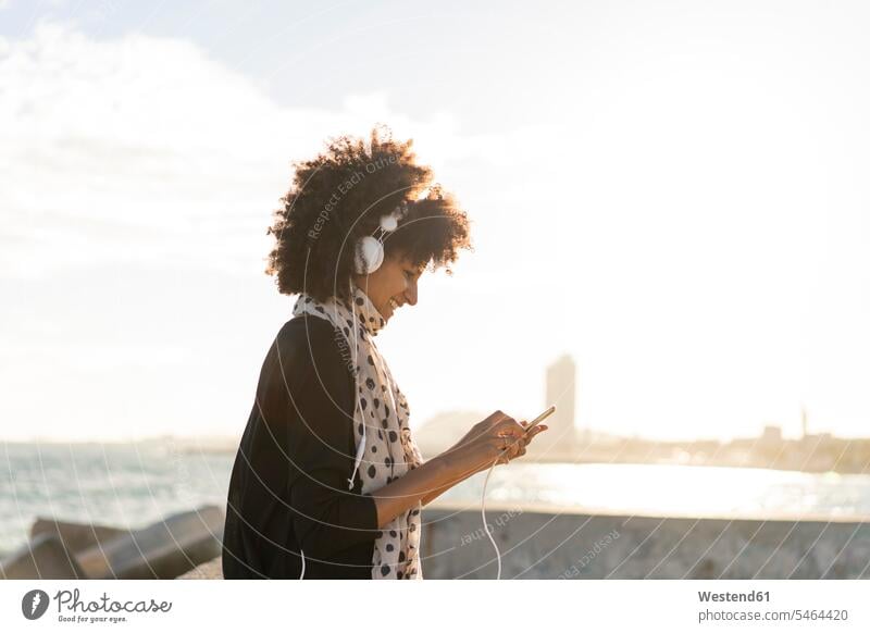 Lächelnde Frau hört Musik mit Smartphone und Kopfhörer in der Nähe des Meeres lächeln hören hoeren benutzen benützen iPhone Smartphones Kopfhoerer weiblich