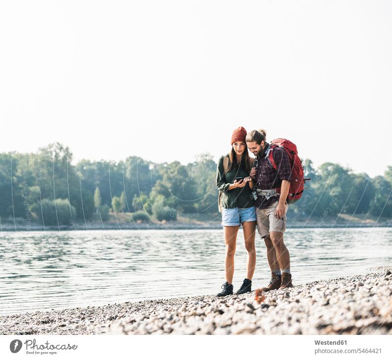 Junges Paar mit Rucksäcken am Flussufer beim Handy-Check Rucksack checken Fluesse Fluß Flüsse Pärchen Paare Partnerschaft Mobiltelefon Handies Handys
