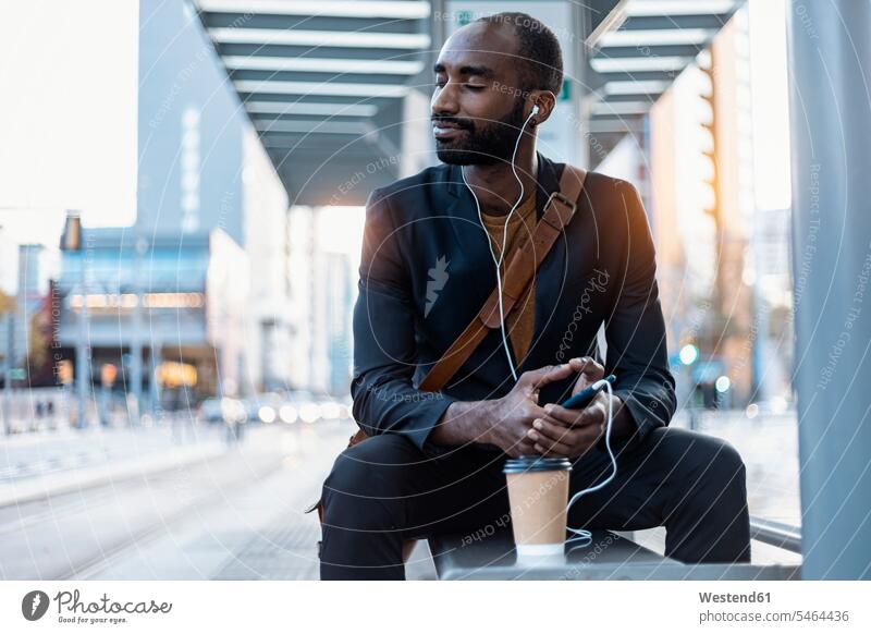 Junger Geschäftsmann mit Kaffee zum Mitnehmen beim Musikhören mit Kopfhörern und Smartphone beim Entspannen an der Straßenbahnhaltestelle Leute Menschen People