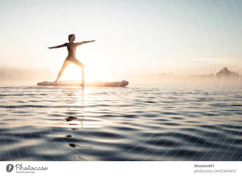Frau praktiziert morgens Paddelbrett-Yoga auf dem Kirchsee, Bad Tölz, Bayern, Deutschland ausüben trainieren Übung früh Frühe Morgen geniessen Genuss stehend