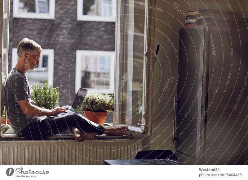Älterer Mann sitzt zu Hause mit Laptop am Fenster Leute Menschen People Person Personen Alleinstehende Alleinstehender Singles Unverheiratete Unverheirateter