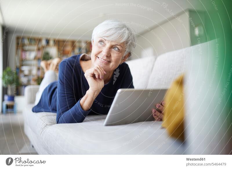 Ältere Frau mit Hand am Kinn, die ein digitales Tablett benutzt, während sie zu Hause auf dem Sofa liegt Farbaufnahme Farbe Farbfoto Farbphoto Innenaufnahme