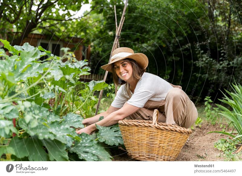Lächelnde junge Frau mit Hut bei der Arbeit im Gemüsegarten während der Ausgangssperre Farbaufnahme Farbe Farbfoto Farbphoto Spanien Latzhose Außenaufnahme