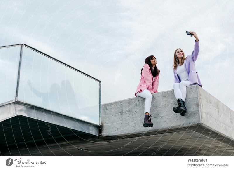 Schwestern nehmen Selfie durch Smartphone, während auf dem Dach in der Stadt gegen den Himmel sitzen Farbaufnahme Farbe Farbfoto Farbphoto Außenaufnahme außen