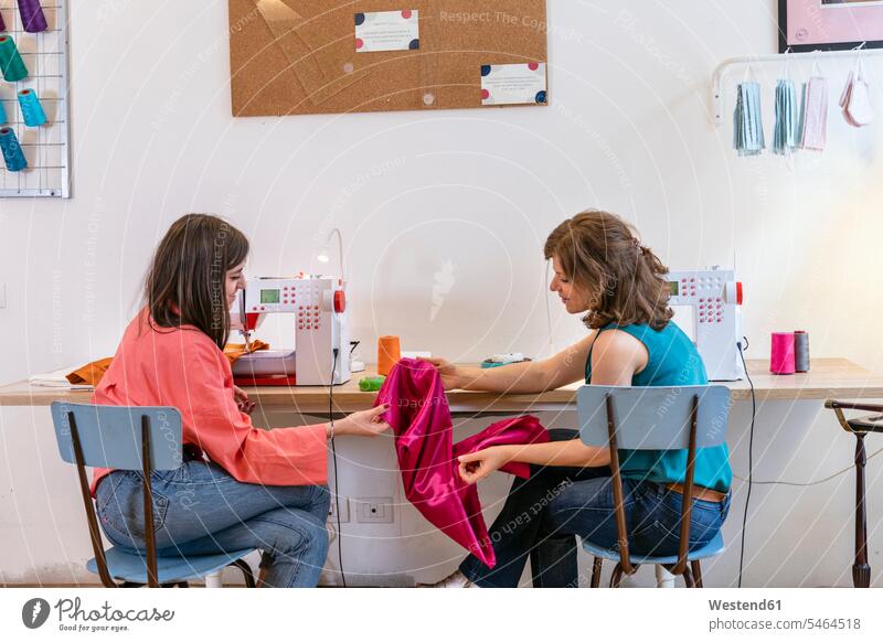 Weibliche Näherin diskutiert über rosa Stoff, während sie im Atelier an Nähmaschinen sitzt Farbaufnahme Farbe Farbfoto Farbphoto Innenaufnahme Innenaufnahmen