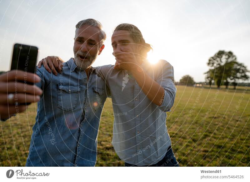 Aufgeregter Vater und erwachsener Sohn schauen sich auf einer Wiese auf dem Land ein Smartphone an Leute Menschen People Person Personen Europäisch Kaukasier