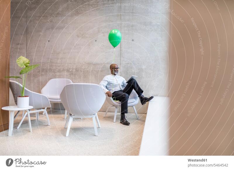 Älterer Geschäftsmann mit grünem Ballon sitzt auf einem Sessel und benutzt ein digitales Tablet Businessmann Businessmänner Geschäftsmänner sitzen sitzend