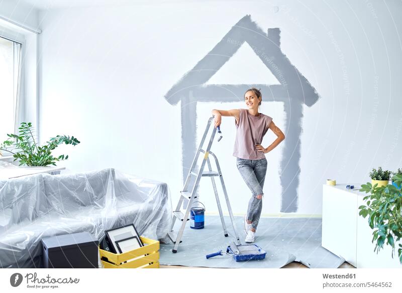 Lächelnde Frau mit Hand auf der Hüfte, die zu Hause gegen ein an die Wand gemaltes Haus steht Farbaufnahme Farbe Farbfoto Farbphoto Innenaufnahme Innenaufnahmen