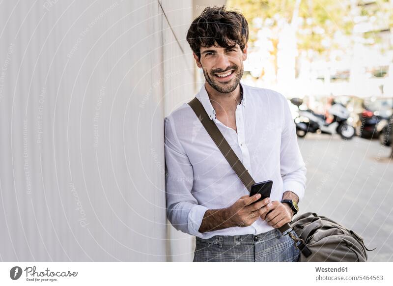 Porträt eines lächelnden jungen Mannes mit Tasche und Mobiltelefon in der Stadt Handy Handies Handys Mobiltelefone Portrait Porträts Portraits staedtisch