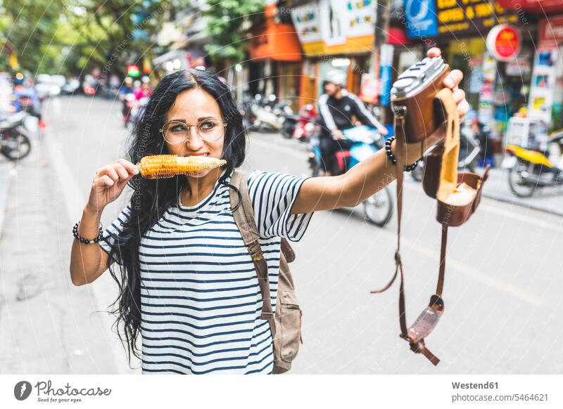 Vietnam, Hanoi, junge Frau macht ein Selfie mit altmodischer Kamera auf der Straße und isst einen Maiskolben Portrait Porträts Portraits Selfies Strassen
