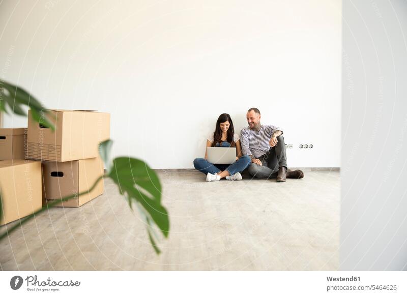 Mann sitzt mit seiner Frau mit Laptop auf Hartholzboden an der Wand in neuem unmöblierten Haus Farbaufnahme Farbe Farbfoto Farbphoto Deutschland Innenaufnahme