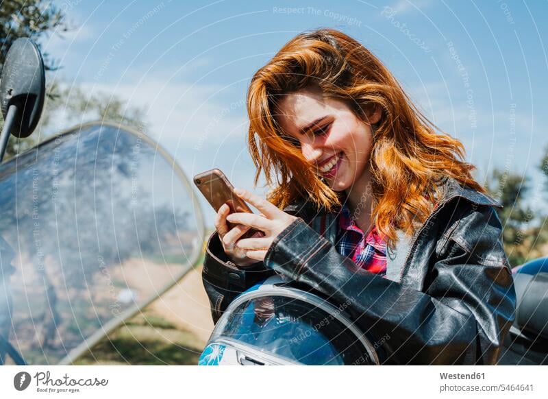 Porträt einer glücklichen rothaarigen Frau auf Motorrad mit Blick auf Handy, Andalusien, Spanien Informationen KFZ Verkehrsmittel Motorräder Telekommunikation