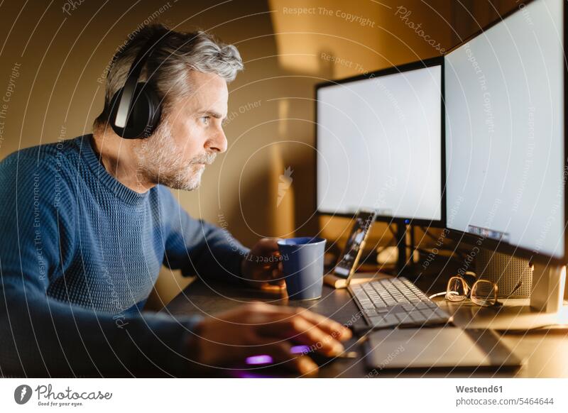 Älterer Mann mit Kopfhörern sitzt zu Hause am Schreibtisch und arbeitet am Computer Job Berufe Berufstätigkeit Beschäftigung Jobs Tische Arbeitstisch