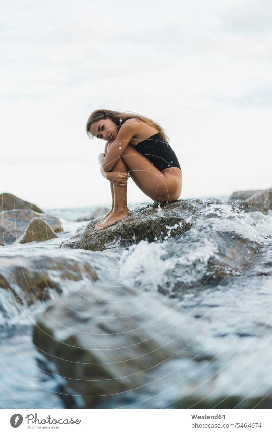 Schöne junge Frau trägt Badeanzug sitzt auf Felsen im Meer Badeanzuege Badeanzüge schön Meere weiblich Frauen sitzen sitzend Badekleidung Badebekleidung