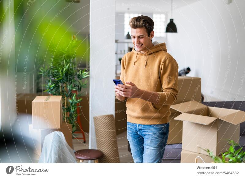 Lächelnder junger Mann, der ein Mobiltelefon in einem unordentlichen Wohnzimmer während eines Umzugs benutzt Farbaufnahme Farbe Farbfoto Farbphoto Innenaufnahme