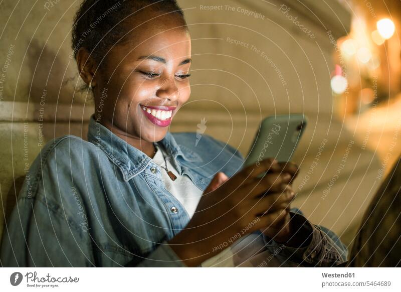 Porträt einer glücklichen jungen Frau, die ein Smartphone in der Stadt bei Nacht benutzt, Lissabon, Portugal Leute Menschen People Person Personen Afrikanisch