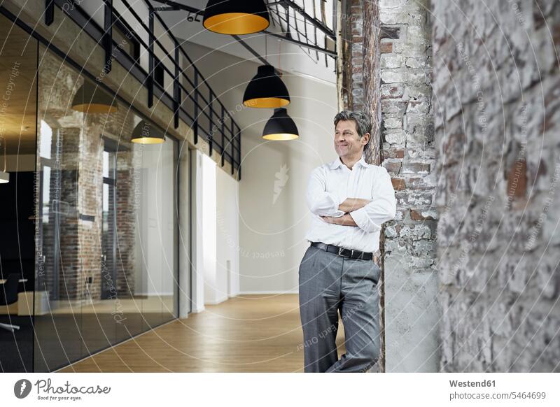 Porträt eines lächelnden Geschäftsmannes, der sich in einem modernen Büro an eine Ziegelmauer lehnt Office Büros Backsteinwand Backsteinmauern Businessmann