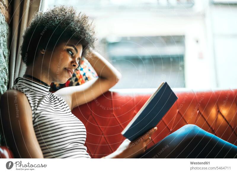 Mittlere erwachsene Frau mit lockigem Haar beim Lesen eines Buches, während sie sich auf dem Sofa im Café entspannt Farbaufnahme Farbe Farbfoto Farbphoto