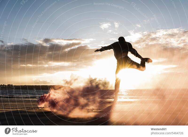 Mann macht Bewegungstraining am Strand mit buntem Rauch bei Sonnenuntergang Qualm Rauchfackel sportlich Training trainieren sich bewegen Beach Straende Strände