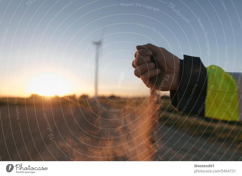 Nahaufnahme der Hand eines Mannes, der bei Sonnenuntergang Erde auf eine Windkraftanlage streut Windturbine Windrad Windräder Hände Feld Felder verstreuen
