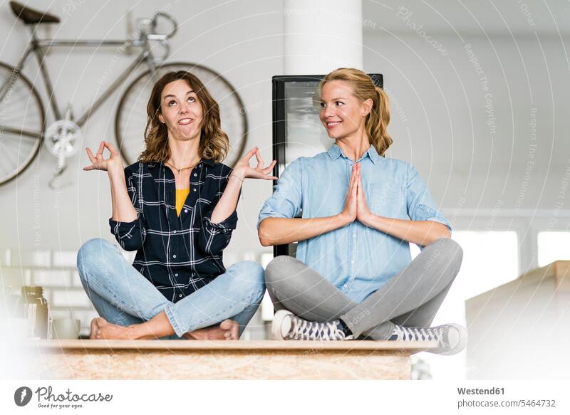 Zwei junge Frauen sitzen im Schneidersitz auf der Theke ihres Cafés, eine meditiert, die andere zieht lustige Gesichter Leute Menschen People Person Personen