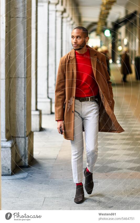 Modischer junger Mann mit rotem Pullover und braunem Mantel, der durch eine Passage geht Arkade Arkaden modisch Männer männlich Mäntel Jacke gehen gehend roter