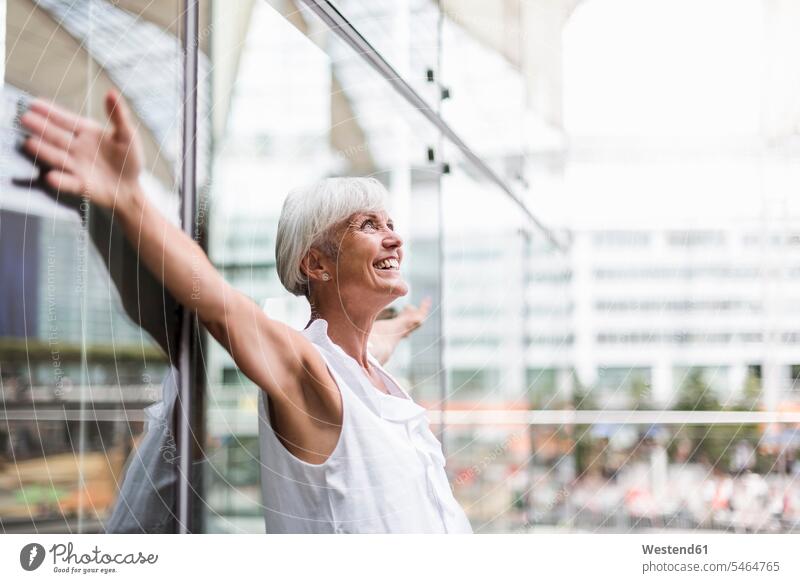 Glückliche ältere Frau lehnt mit ausgestreckten Armen an einer Glasfassade anlehnen angelehnt lehnend Seniorin Seniorinnen alt Portrait Porträts Portraits