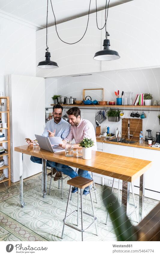 Zwei junge Männer arbeiten zusammen in der Küche, benutzen einen Laptop, diskutieren Dokumente Arbeit Unterlagen Küchen Freunde besprechen Besprechung