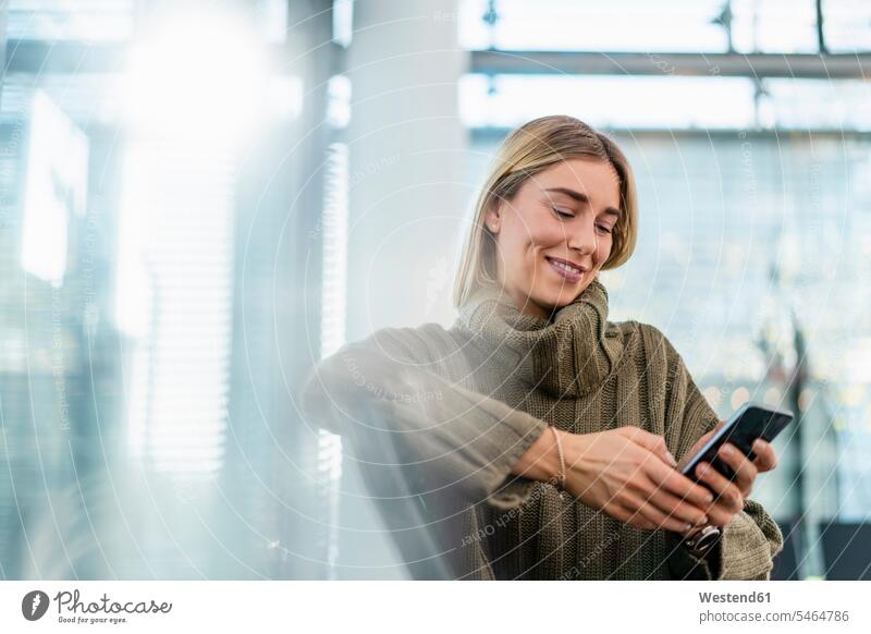 Lächelnde junge Frau sitzt im Wartebereich und benutzt ein Mobiltelefon Informationen Telekommunikation telefonieren Handies Handys Mobiltelefone verreisen