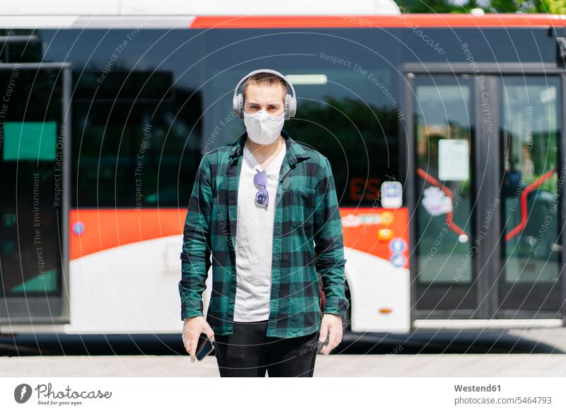 Porträt eines jungen Mannes mit Schutzmaske, der vor dem Bus mit Kopfhörern Musik hört, Spanien Transport Transportwesen KFZ Verkehrsmittel Autobus Autobusse