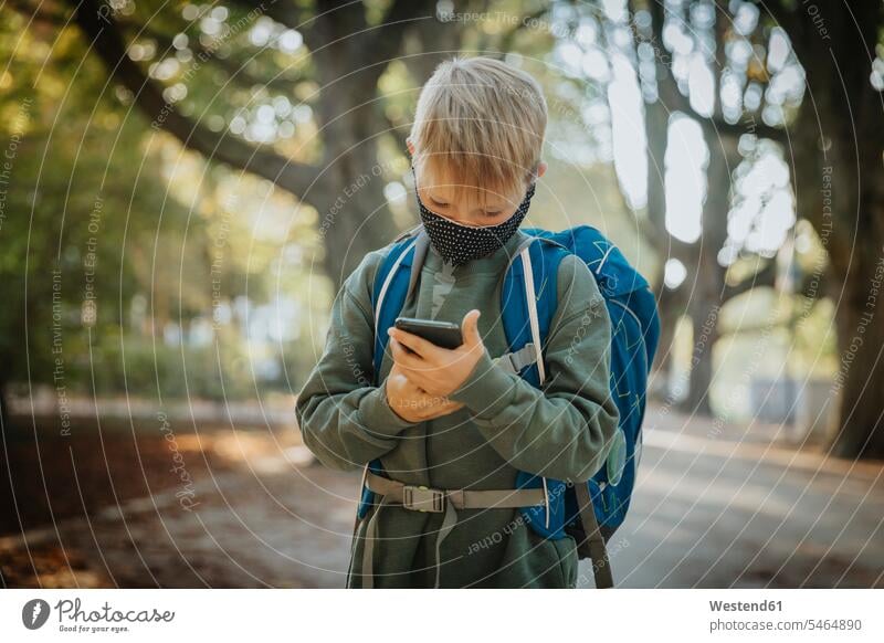 Textnachrichten von Jungen mit Smartphones, die eine schützende Gesichtsmaske tragen, wenn sie im öffentlichen Park stehen Farbaufnahme Farbe Farbfoto Farbphoto