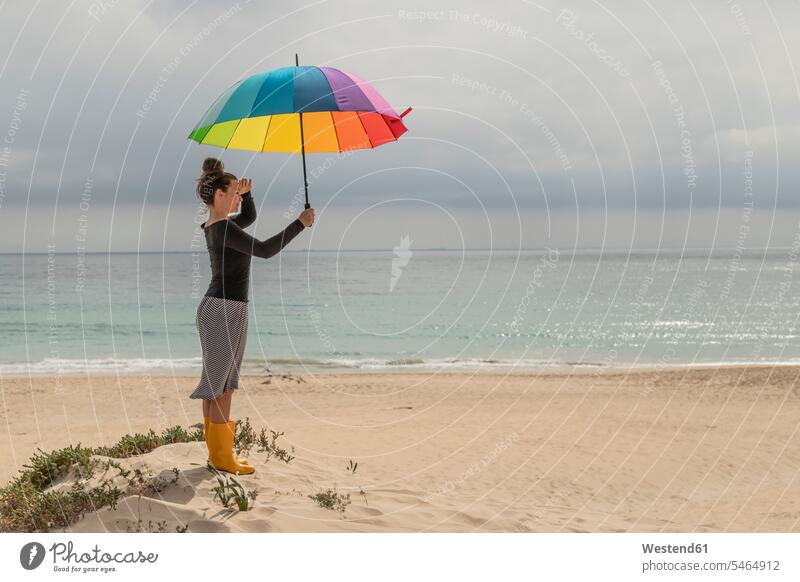 Frau mit bunten Regenschirm sitzt am Strand Regenbogenfarben Spektralfarben Toleranz tolerant Gummistiefel Ausschau halten weiblich Frauen multikulturell Beach