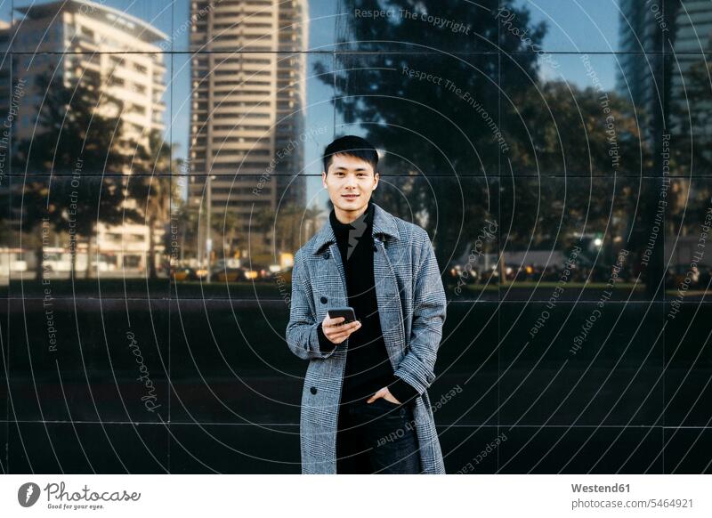 Spanien, Barcelona, Porträt eines jungen Mannes mit Mobiltelefon, der einen schwarzen Rollkragenpullover und einen grauen Mantel trägt Portrait Porträts