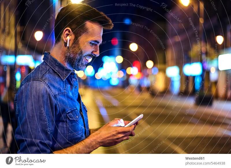 Mann mit drahtlosen Kopfhörern mit Smartphone in der Stadt bei Nacht Leute Menschen People Person Personen Europäisch Kaukasier kaukasisch 1 Ein ein Mensch eine