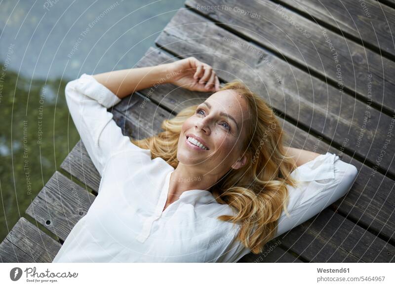 Lächelnde blonde Frau auf Holzsteg an einem See liegend blonde Haare blondes Haar hölzern Steg Stege Anlegestelle liegt weiblich Frauen Seen lächeln Mensch