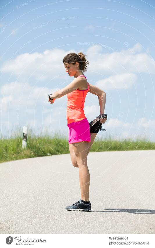 Lächelnde junge Frau streckt ihr Bein und schaut auf die Smartwatch Joggerin Joggerinnen trainieren Dehnübung dehnen strecken ansehen weiblich Frauen Joggen