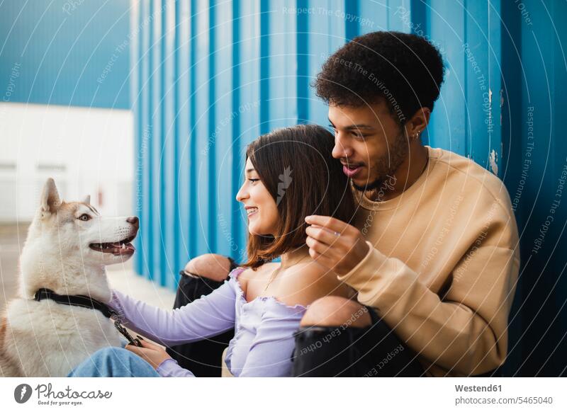 Lächelndes Paar sitzt mit Hund und lehnt sich an blaue Wand Farbaufnahme Farbe Farbfoto Farbphoto Außenaufnahme außen draußen im Freien Tag Tageslichtaufnahme
