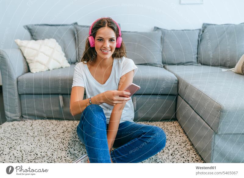 Porträt einer lächelnden jungen Frau, die im Wohnzimmer auf dem Boden sitzt und mit einem Mobiltelefon Musik hört Leute Menschen People Person Personen