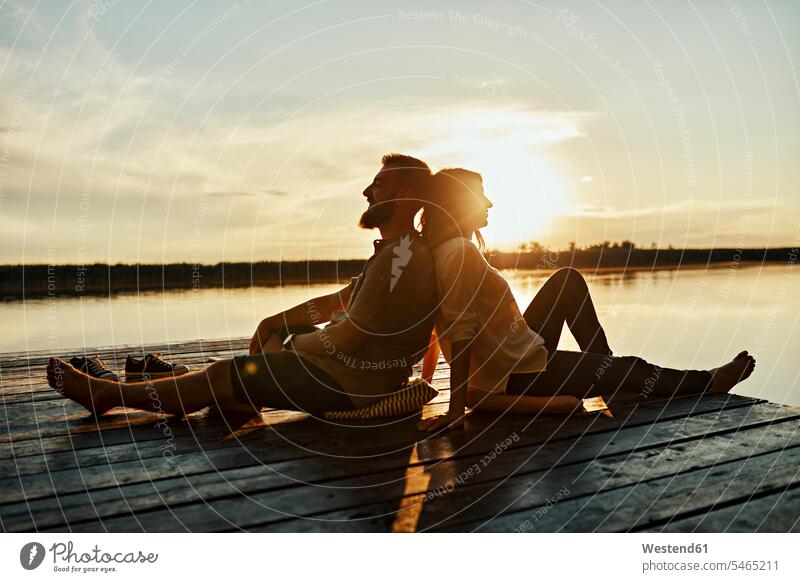 Paar sitzt Rücken an Rücken auf einem Steg an einem See bei Sonnenuntergang Polster sitzend abends Jahreszeiten sommerlich Sommerzeit entspannen relaxen