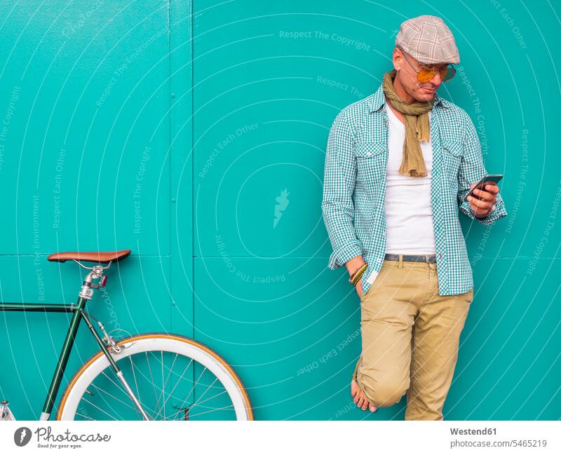 Mann mit Fixie-Fahrrad steht vor grüner Wand und schaut auf Handy Informationen Schals Raeder Räder Bike Bikes Fahrräder Rad Telekommunikation Handies Handys