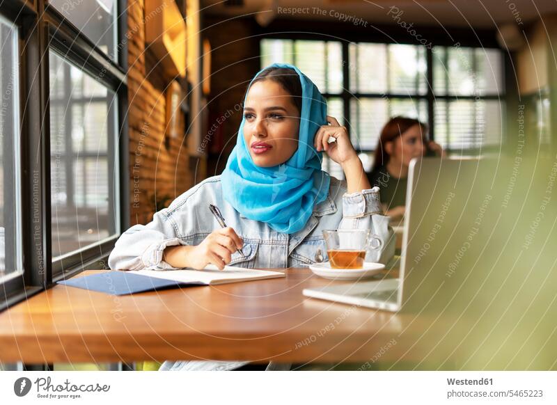 Geschäftsfrau, die in einem Café einen türkisfarbenen Hidschab trägt und in ein Notizbuch schreibt Leute Menschen People Person Personen Europäisch Kaukasier