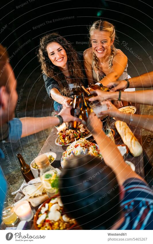 Glückliche Freunde stoßen bei einem Abendessen im Freien mit Bierflaschen an Kameradschaft Flaschen Tische entspannen relaxen abends nachts Jahreszeiten