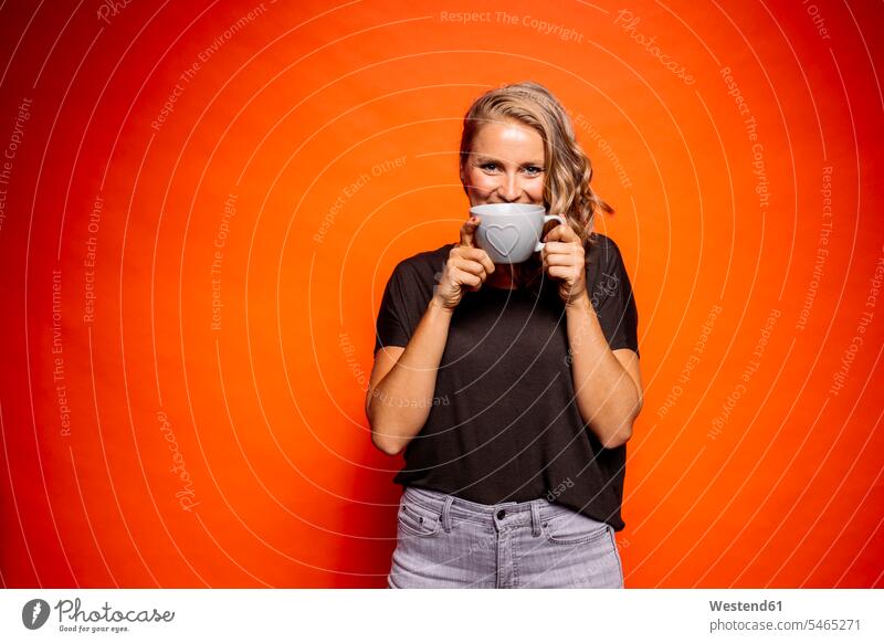 Glückliche Frau hält Kaffeetasse mit Herzform in der Hand, während sie vor orangem Hintergrund steht Farbaufnahme Farbe Farbfoto Farbphoto farbiger Hintergrund