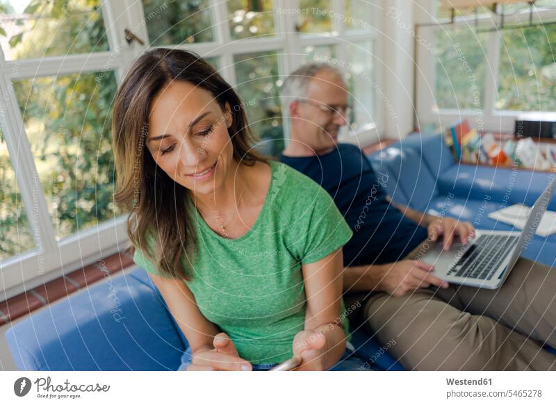 Reifes Paar sitzt auf Couch zu Hause mit Frau mit Handy und Mann mit Laptop Couches Liege Sofas daheim sitzend Rechner Laptops Notebook Notebooks
