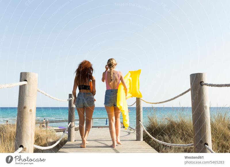 Rückansicht von jungen Frauen mit gelber Luftmatratze und Sonnenschirm, die zum Strand gehen Luftmatratzen gehend geht sommerlich Sommerzeit frei Muße