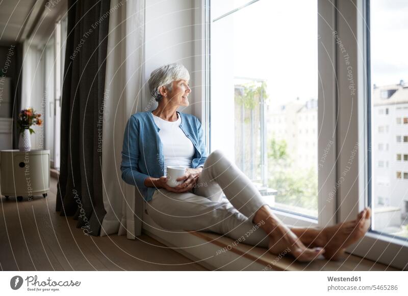 Lächelnde ältere Frau, die zu Hause sitzend durchs Fenster schaut Farbaufnahme Farbe Farbfoto Farbphoto Innenaufnahme Innenaufnahmen innen drinnen Tag