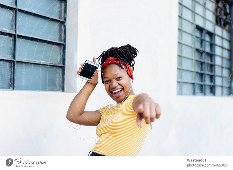 Porträt einer aufgeregten jungen Frau, die mit Kopf- und Ohrhörern vor einer weißen Wand Musik hört Leute Menschen People Person Personen Afrikanisch