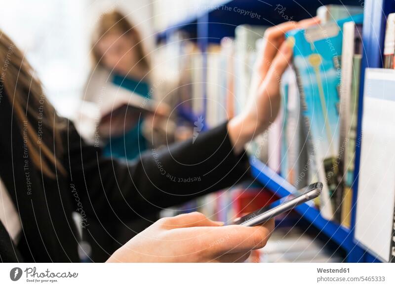 Hand einer Teenagerin, die ein Handy hält, während sie ein Buch aus einem Regal in einer öffentlichen Bibliothek nimmt Smartphone halten Bücherei nehmen
