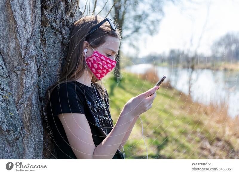 Mädchen mit selbstgemachter Schutzmaske, das ein Smartphone benutzt und sich an einen Baum lehnt Telekommunikation telefonieren Handies Handys Mobiltelefon